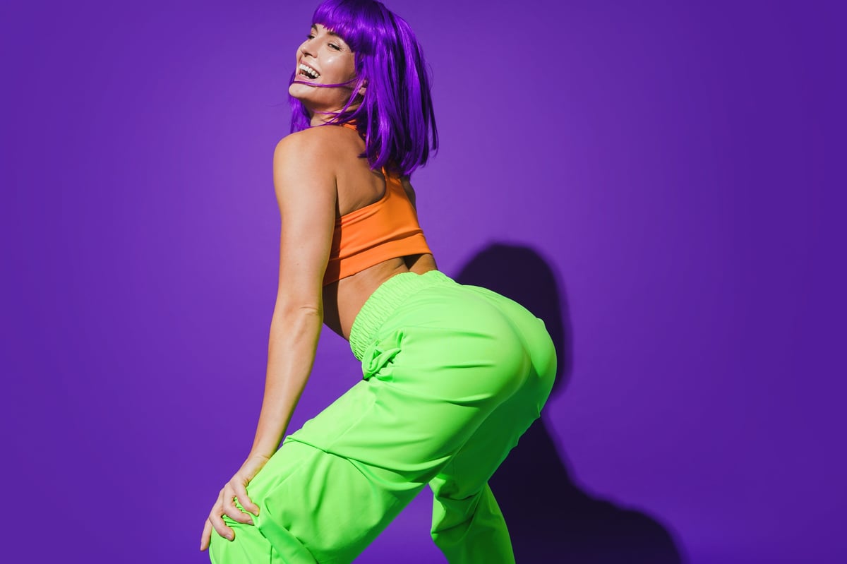 Carefree woman dancer wearing colorful sportswear twerking against purple background