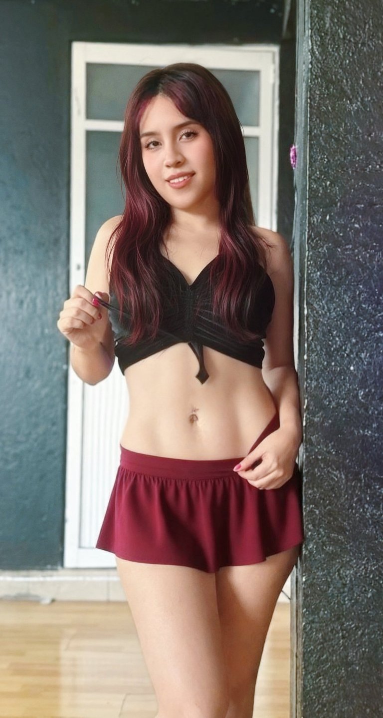 Young woman with long burgundy hair wearing a black crop top and maroon skirt, standing indoors near a white-framed window